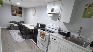 Een moderne keuken met witte kasten, een magnetron, fornuis en gootsteen. Er is een eethoek met een tafel en stoelen.