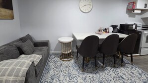 Een knus woongedeelte met een bank, een eettafel met stoelen, een klok, een magnetron en een koffiezetapparaat.