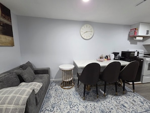 A compact living space with a sofa, a dining table with chairs, a clock, a microwave, and a coffee maker.