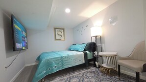 Een slaapkamer met een bed, een stoel, een klein tafeltje, een vloerlamp, een tv aan de muur en een kamerkleed met dessin.