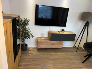 A modern living room with a flat-screen TV mounted on the wall, a wooden cabinet with a plant, and a small table with a remote control.