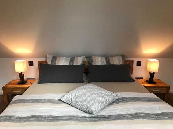 Ein ordentlich gemachtes Bett mit einem gemusterten Bettüberwurf, zwei dunklen Kissen und einem geometrischen Kissen. Es gibt zwei Nachttische mit Lampen, jeder mit einem hölzernen Fuß und einem kleinen Bilderrahmen.