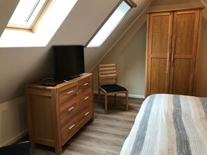 Ein Schlafzimmer mit schrägem Dach, einem Holzschrank, einem Fernseher auf einer Kommode, einem Sessel und einem Bett.
