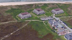 Oversigtsbillede af en ejendom ved kysten med tre store bygninger, et parkeringsområde og en strand i baggrunden.