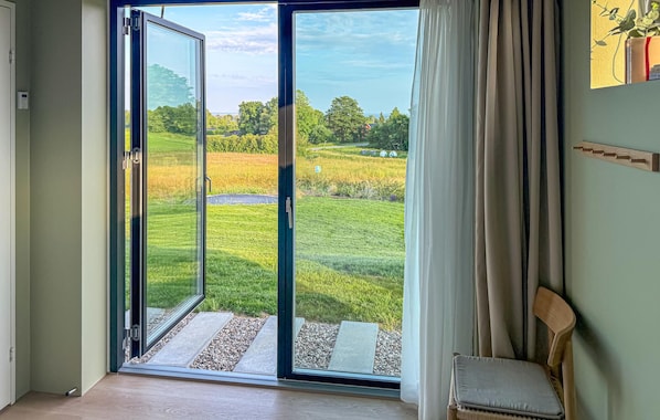 A room with a sliding glass door leading to a grassy outdoor area, a chair, and a wall-mounted shelf with a plant. - 4 bedroom beautiful home in Haderslev (Haderslev)