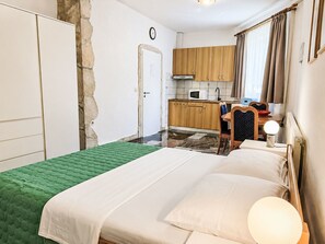 Un dormitorio con cama, colcha verde, una silla de madera, un escritorio con lámpara, una zona de cocina con armarios y un microondas, y una pared de piedra.