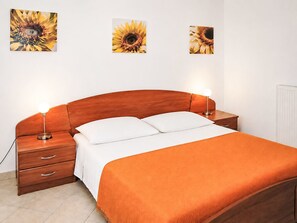 Ein Schlafzimmer mit einem Holz-Kopfteil, zwei Nachttischen, einem Bett mit weißer und orangefarbener Bettwäsche sowie drei Sonnenblumen-Wandbildern.