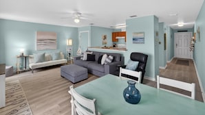 A living room with a grey sofa, a white armchair, a dining table with a vase, and a kitchen area in the background. - Y Knot Go to the Beach! Cute Beach Condo (Corpus Christi)