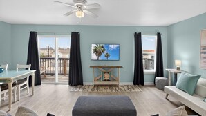 A living room with a sofa, ottoman, and a dining table with chairs. There is a ceiling fan, a TV on the wall, and a window with curtains. - Y Knot Go to the Beach! Cute Beach Condo (Corpus Christi)