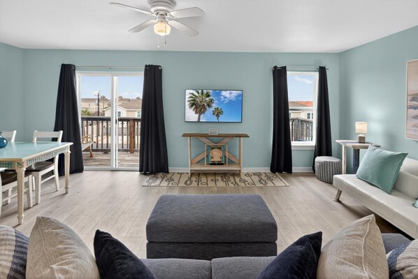 Un salon comprenant un canapé, un pouf et une table à manger avec des chaises. On y trouve également un ventilateur de plafond, un téléviseur fixé au mur et une fenêtre avec des rideaux.
