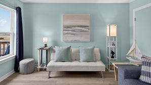 A room with a light blue wall, a white sofa with light blue cushions, a small wooden side table with a lamp, a floor lamp, and a view of the outdoors through a window. - Y Knot Go to the Beach! Cute Beach Condo (Corpus Christi)