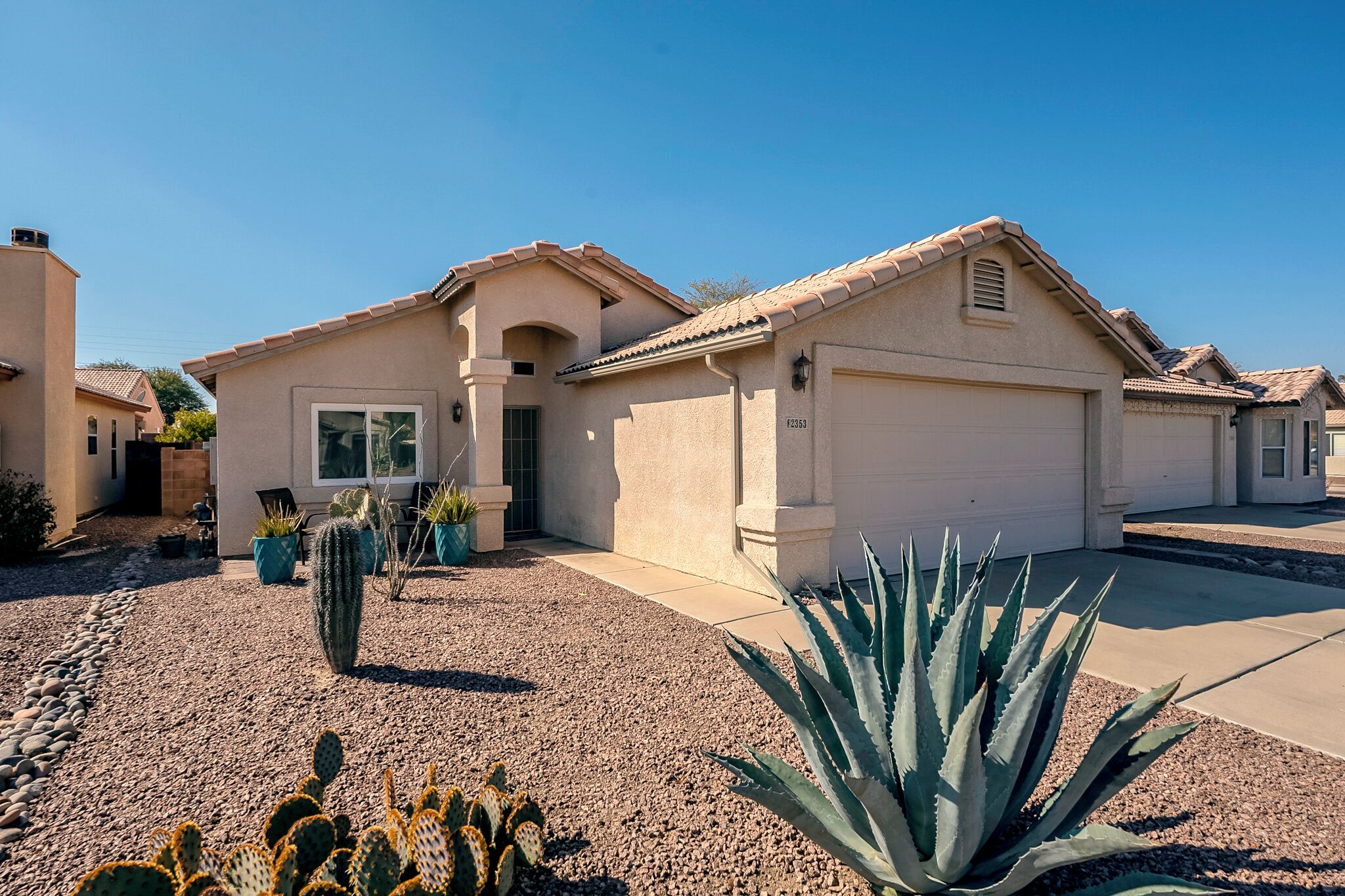 Une maison d’habitation avec un garage, une porte d’entrée et un petit patio agrémenté de cactus et d’une grande plante d’agave.
