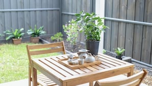 Une table en bois, sur laquelle sont disposés un plateau de tasses à thé et une théière, est placée à l’extérieur, avec une clôture en bois et des plantes en pot en arrière-plan.
