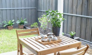 Une table en bois, sur laquelle sont disposés un plateau de tasses à thé et une théière, est placée à l’extérieur, avec une clôture en bois et des plantes en pot en arrière-plan.