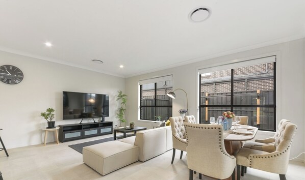A modern living room with a flat-screen TV, a sofa, a dining area with chairs, and a large window with a view of a brick building. - KozyGuru | Box Hill | New Designer Home (Box Hill)