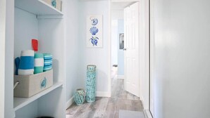 A hallway with light blue walls, a wooden floor, and a door leading to another room. Shelves on the left hold folded towels and a decorative item. - The HotTub Hideaway-5 Min From Sombrero Beach (Marathon)