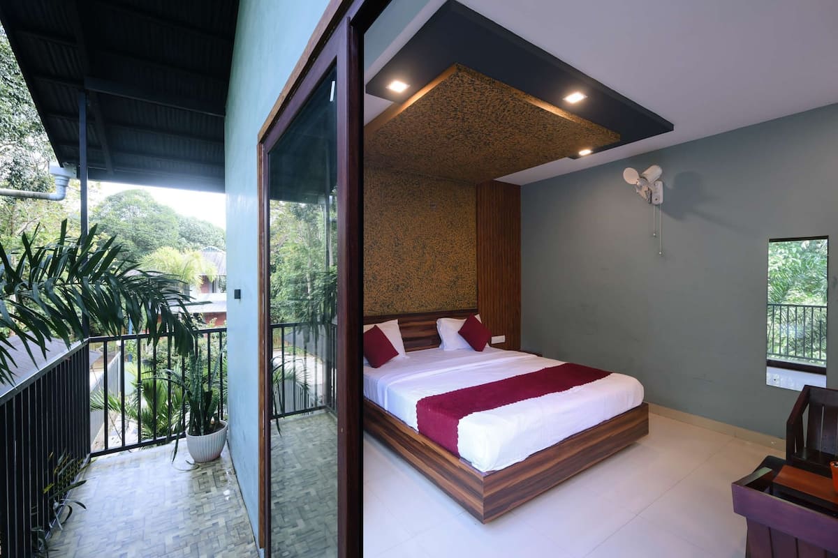 A modern bedroom with a large bed, a balcony with potted plants, and a view of greenery.