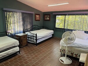 A room with two beds, a bedside table, a fan, and a window with blinds.