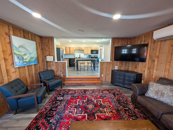 A living room with a large rug, a sofa, a TV, and a kitchen in the background.