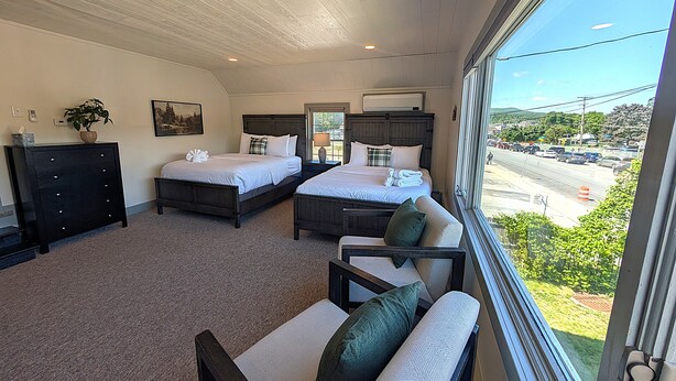 A hotel room with two beds, a sofa, a dresser, a plant, and a large window with a view of the street. - Village Overlook: Spacious 5 BR + Prime Location (Lake George)