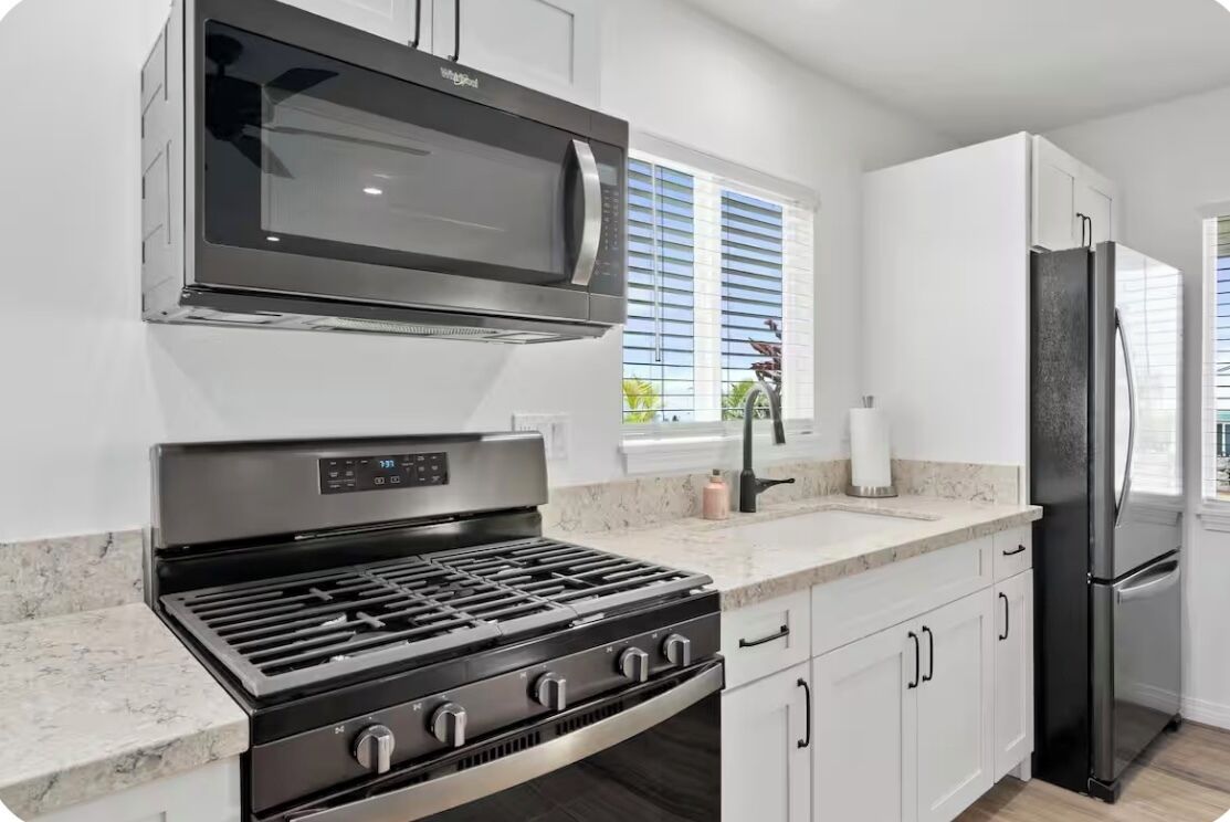A modern kitchen with a stainless steel range, microwave, and refrigerator, white cabinetry, and a window with blinds.