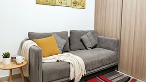 Ein graues Sofa mit gemusterten Kissen, ein kleiner runder Holztisch mit einer Pflanze und einer weißen Tasse sowie ein rot-schwarzer Streifen Teppich.