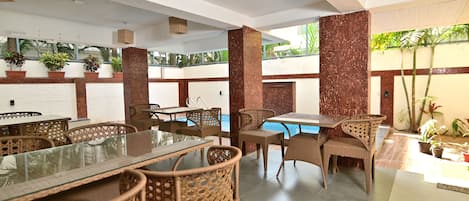 Un comedor interior con mobiliario de mimbre, una mesa con superficie de vidrio y una piscina al fondo.