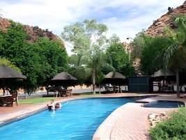 Una piscina con áreas de descanso alrededor, árboles y montañas al fondo.