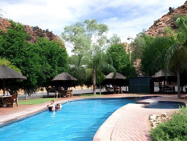 A swimming pool with surrounding lounge areas, trees, and a mountainous backdrop.