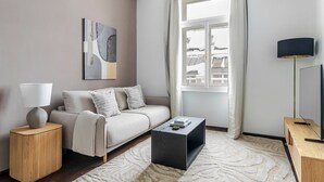 Un salon moderne avec un canapé, une table basse, une lampe, un tableau et une vue sur des immeubles par la fenêtre.