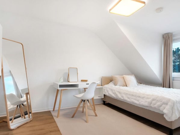 A modern bedroom with a sloped ceiling, a bed with white bedding, a desk with a chair, and a large mirror.