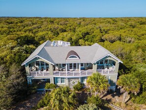 Une grande maison à deux étages, dotée d’un balcon et de nombreuses fenêtres, et entourée d’une végétation luxuriante.