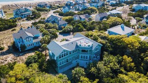 Luchtbeeld van een kustgebied met woonhuizen, voornamelijk blauw, sommige met grote ramen en balkons, omringd door groen en een strand op de achtergrond.
