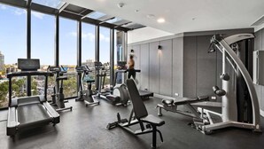 Ett gym med olika typer av träningsutrustning, stora fönster och utsikt över stadsbilden.