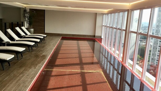 Un espacio interior amplio con suelo de baldosas rojas, una fila de sillones blancos y ventanas grandes con vistas a la ciudad.