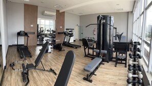 A modern gym with cardio machines, weightlifting equipment, and a large window providing a view of the city. - Qs Marista Stay And Hotel (Goiânia)