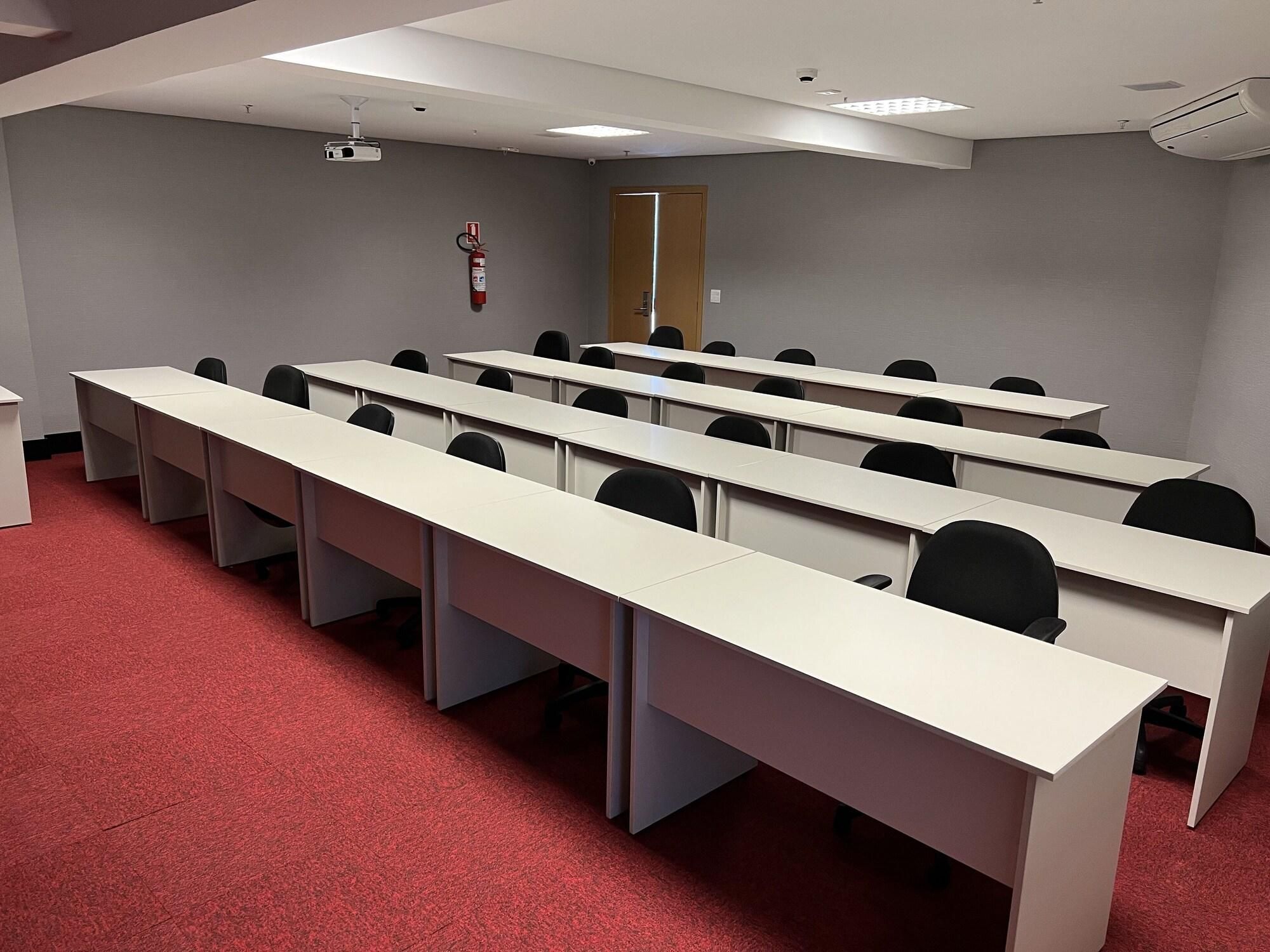 Un salón de conferencias con mesas largas y sillas, un proyector, un extintor y una puerta.