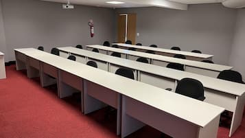 Un salón de conferencias con mesas largas y sillas, un proyector, un extintor y una puerta.