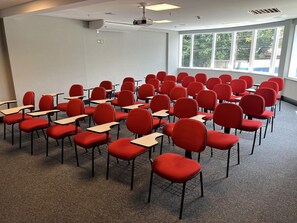 A classroom with rows of red chairs and desks, large windows, and a ceiling fan. - Qs Marista Stay And Hotel (Goiânia)