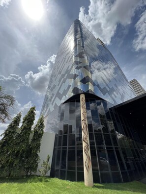 Un edificio moderno con fachada de vidrio reflectante, una columna central destacada con diseño geométrico, y un cielo despejado con nubes.