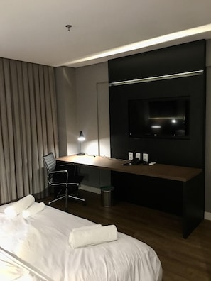 Una habitación de hotel moderna con una cama grande, un escritorio con computadora, una silla, un televisor montado en la pared y una lámpara.