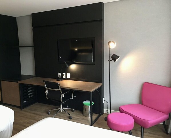 Et hotelværelse med en seng, et skrivebord med stol, et fjernsyn, en gulvlampe og en lyserød sofa.