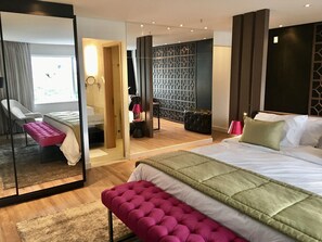 Quarto de hotel moderno com uma cama grande, área de estar com um banco, espelho e vista para o exterior.