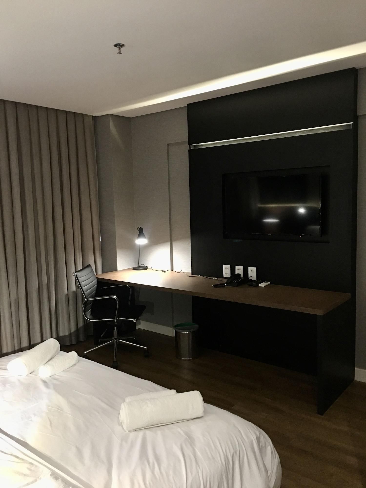 Una habitación de hotel moderna con una cama grande, un escritorio con computadora, una silla, un televisor montado en la pared y una lámpara.
