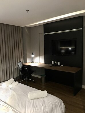 A modern hotel room with a large bed, a desk with a computer, a chair, a TV mounted on the wall, and a lamp. - Qs Marista Stay And Hotel (Goiânia)