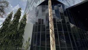 Un edificio moderno con fachada de vidrio reflectante, una columna central destacada con diseño geométrico, y un cielo despejado con nubes.