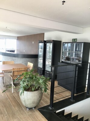 A modern interior with a wooden dining table, chairs, a large potted plant, and two refrigerators with beer labels. - Qs Marista Stay And Hotel (Goiânia)
