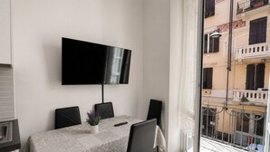 Ett modernt rum med ett matbord, stolar, en tv som är monterad på väggen och en balkong med utsikt över byggnaderna.
