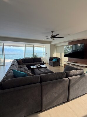 A modern living room with a large sectional sofa, a flat-screen TV mounted on a wooden wall, and a view of the sea through large windows. - 4 BR Luxury oceanfront corner residence at Tres Mares in Marina Vallarta PV (Puerto Vallarta)