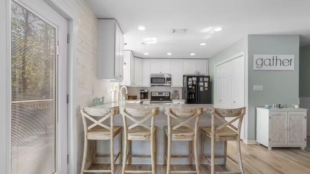 A modern kitchen with white cabinets, a central island with wooden chairs, and a 'gather' sign on the wall. - Coastal Home with Outdoor Oasis. Golf Cart Avail (Long Beach)